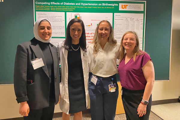 Four female fellows indoors in front of a presentation poster